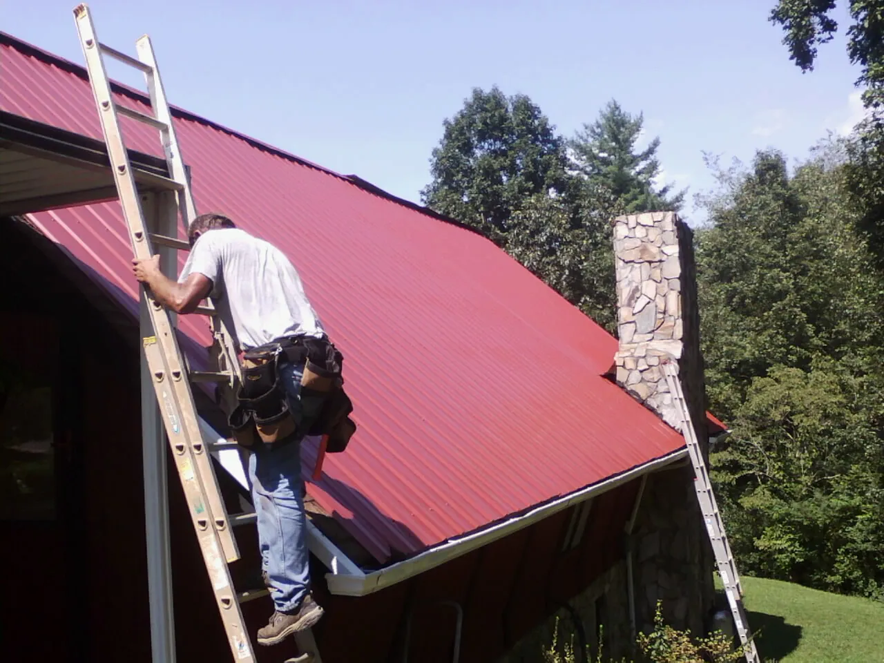 Roofer on a ladder beside a red metal roof and stone chimney — Soffit & Fascia Repair by Roofing Brothers in Margaret